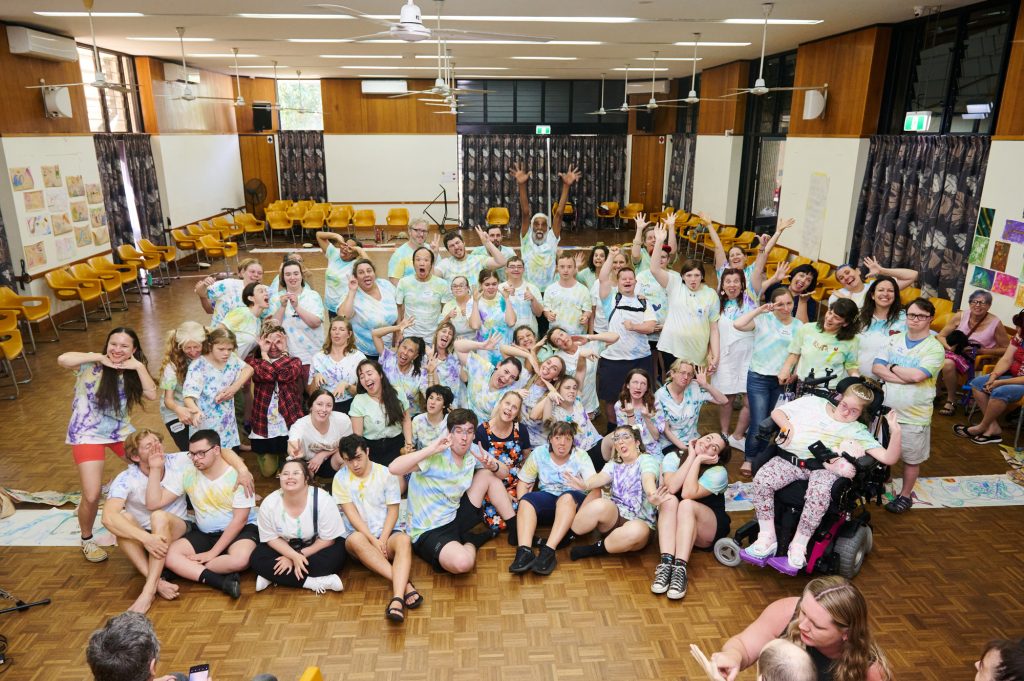A large group of people in matching tie-dyed t-shirts are gathered together to pose for a group photo in a hall with timber floors. Some are seated while others stand and a person at the front uses a motorised wheelchair, many are pulling silly faces or pose in a funny way.