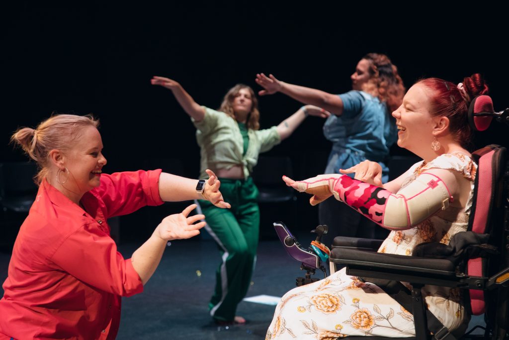 A person with a red shirt and a person with red hair and a white dress and a colourful arm brace, seated in an electronic wheelchair, dance together. In the background, a person wearing green and a person wearing blue are also dancing.