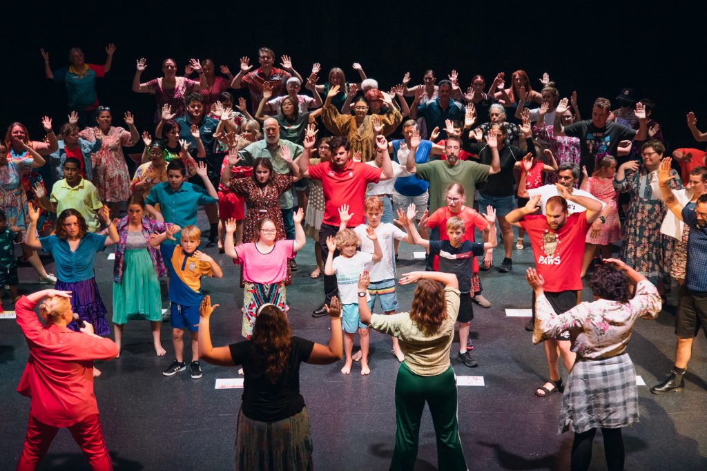 Four people are facing away from the camera, posing with their arms up. Facing them, a large group of people, including children, copy their poses.