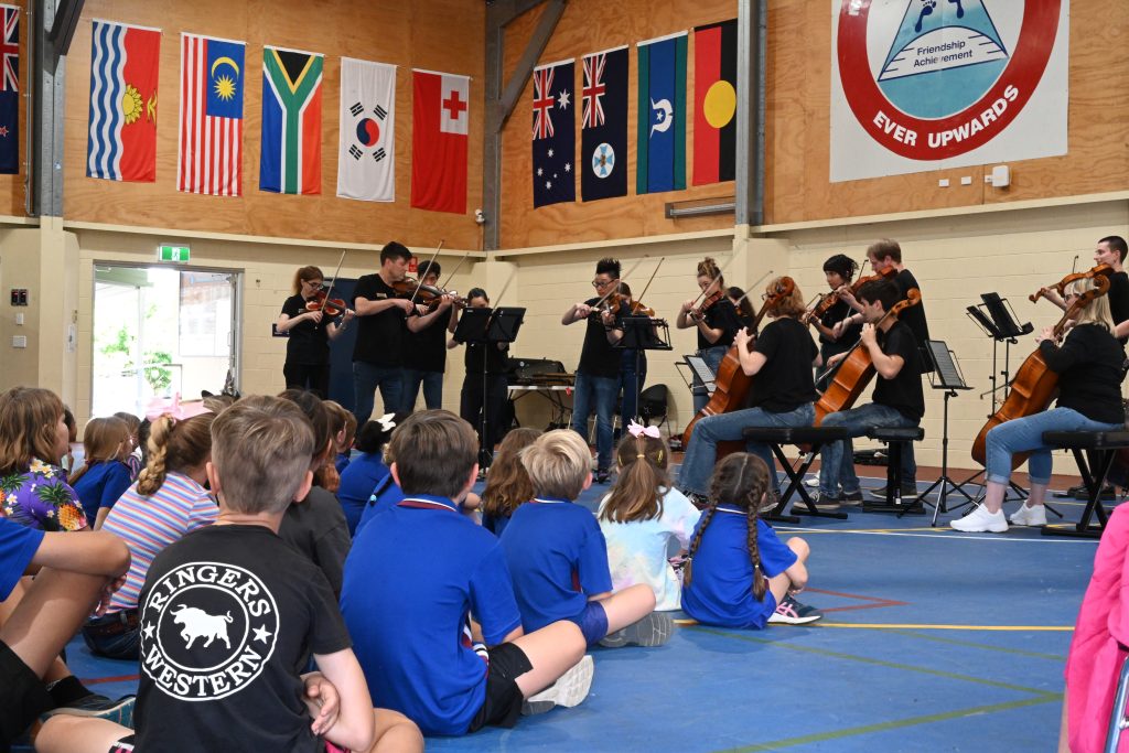 A group of children in school uniforms sit facing away from the camera, watching a group of musicians perform. There are flags of different countries hanging on the wall, and the musicians are playing violins, violas, and cellos.